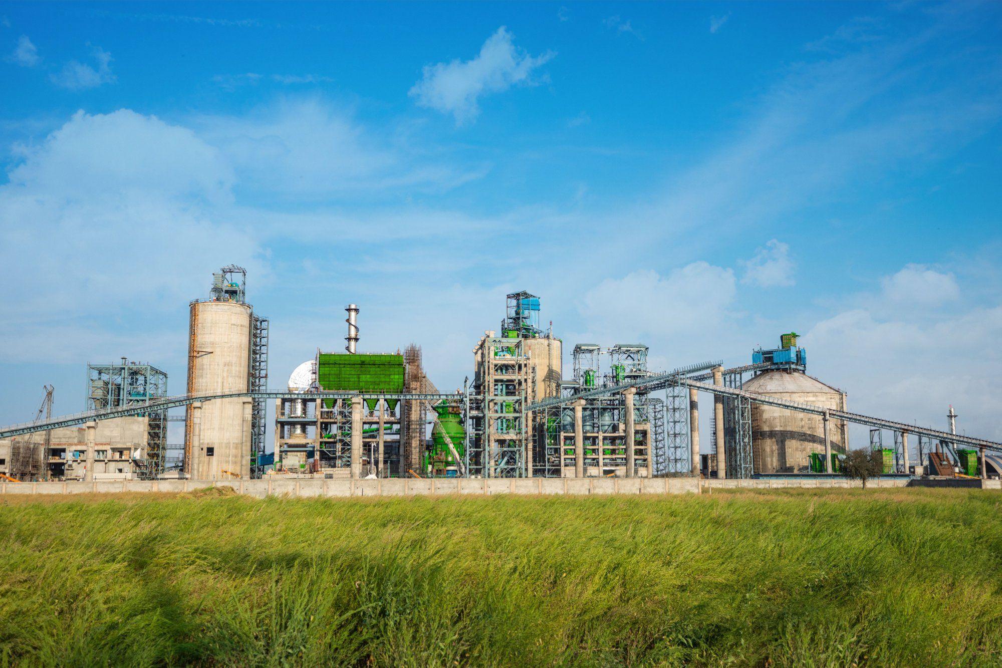 Large industrial plant with various buildings, silos, and green field under a blue sky.