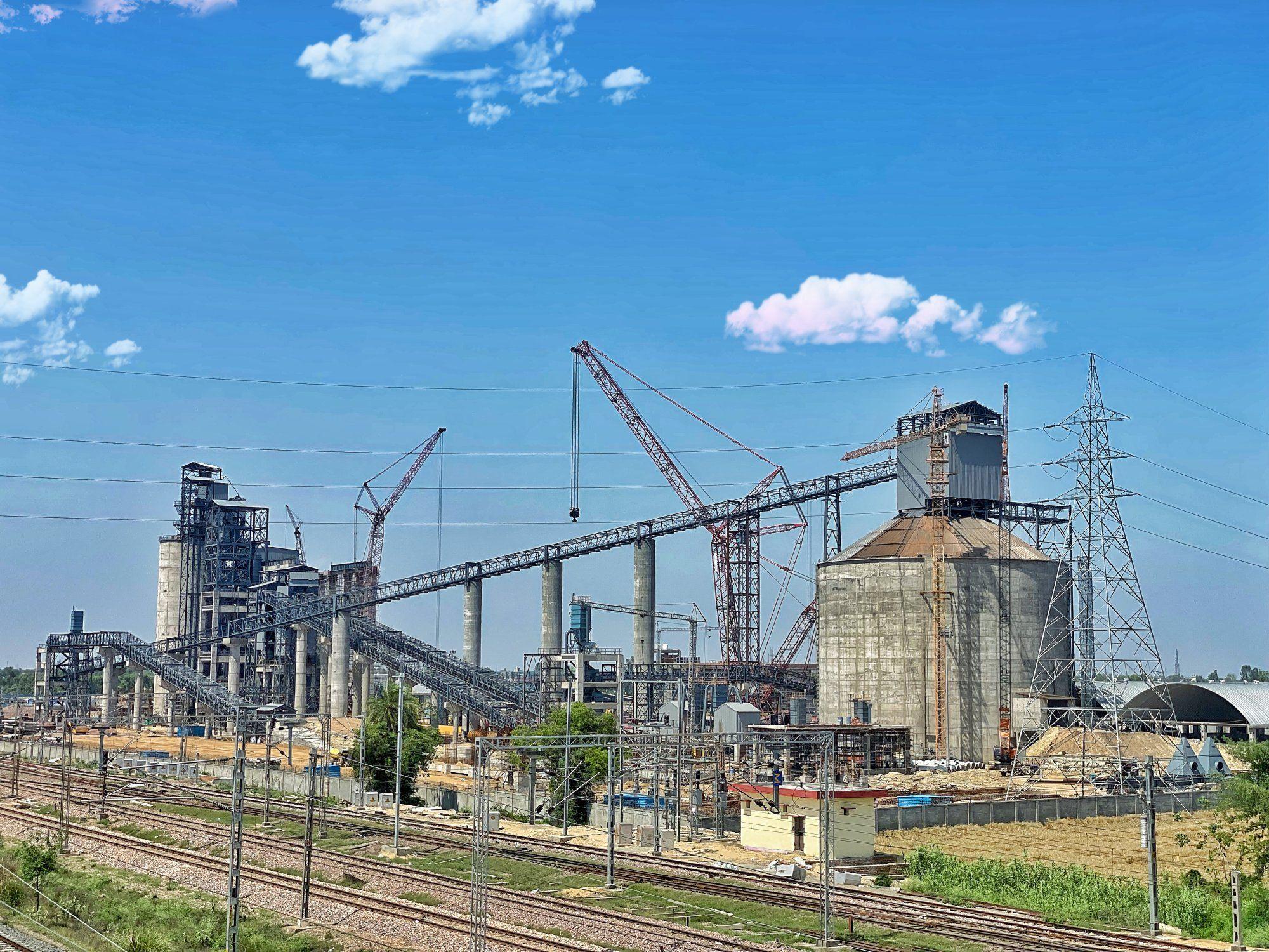 Industrial cement plant featuring large silos, conveyor belts, and cranes beside railway tracks.