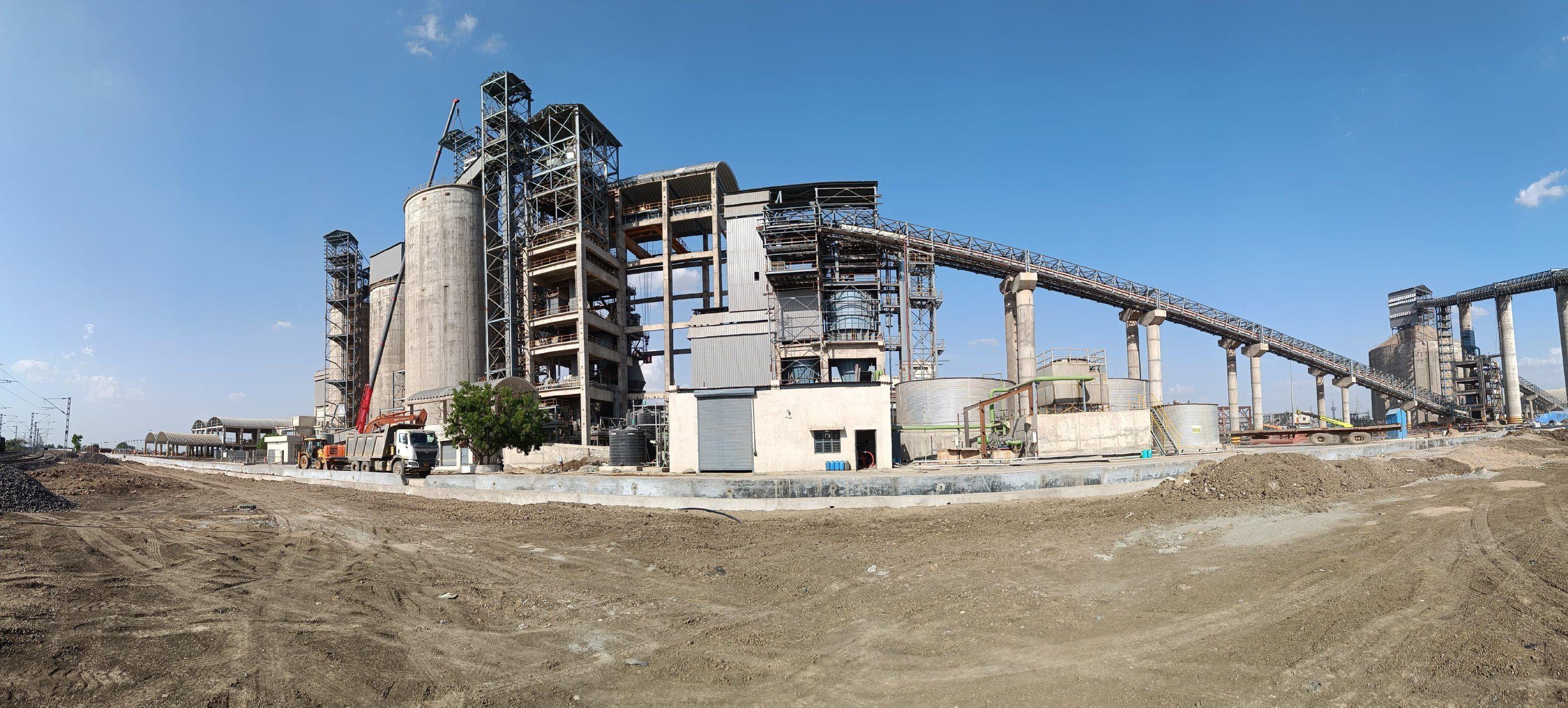 Panoramic view of a large industrial plant with tall silos and conveyor belts under blue sky.