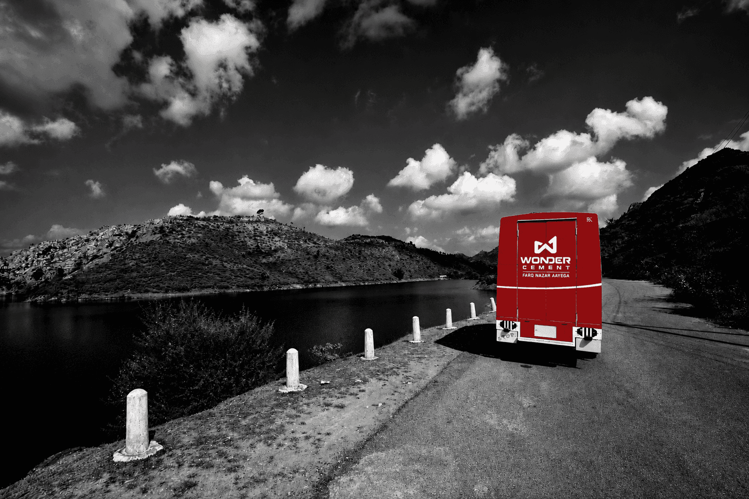 Red truck on winding road by lake, contrasted against a grayscale mountain landscape.