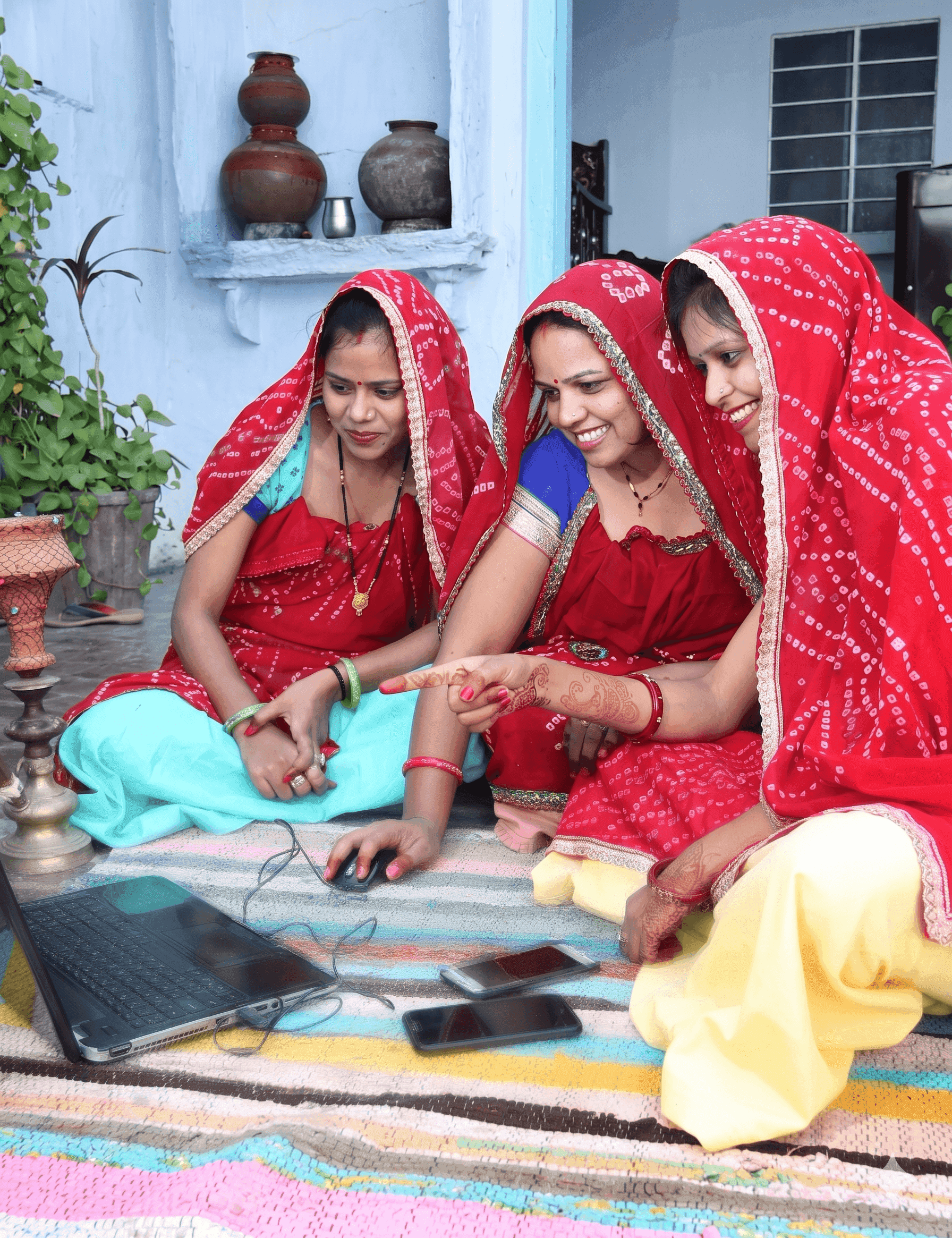 Three Indian women in traditional attire happily engaging with a laptop outdoors.