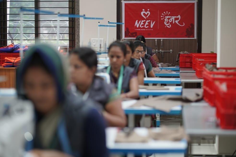 Women work diligently at sewing machines in a bright workshop under a 'NEEV हुनर' banner.