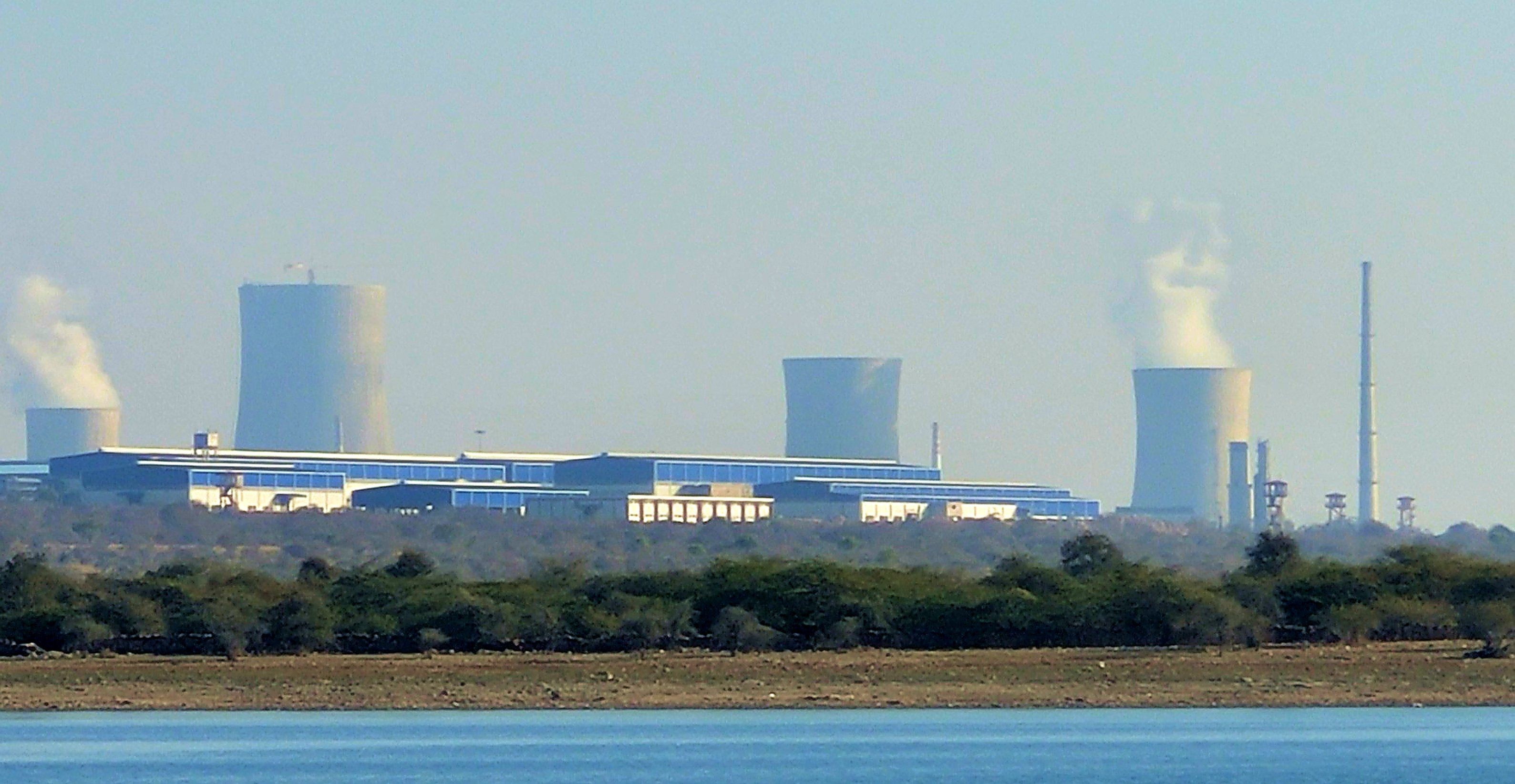 A large power plant with several cooling towers emitting steam, seen across water and trees.